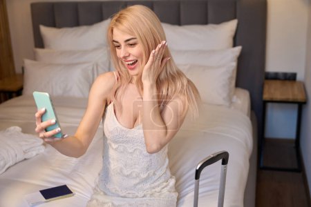 Photo for Excited blond woman keeping video call sitting at double bed in hotel suite, arrived abroad to weekend, bragging with engagement ring - Royalty Free Image