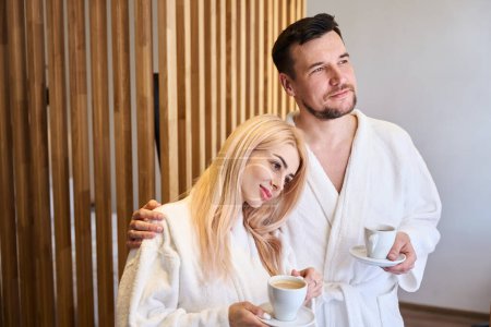 Téléchargez les photos : Couple marié heureux debout dans des peignoirs blancs doux, boire le café du matin et regarder la vue dans la fenêtre, routine quotidienne agréable - en image libre de droit