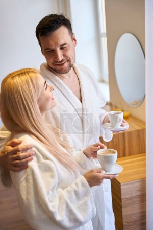 Téléchargez les photos : Couple joyeux se regardant avec amour et tendresse, debout dans des peignoirs chauds et en buvant du café, profiter du week-end - en image libre de droit