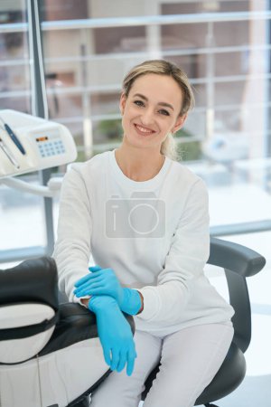Téléchargez les photos : Jeune femme dans des gants de protection assis sur la chaise, se préparer pour le travail dans la stomatologie - en image libre de droit