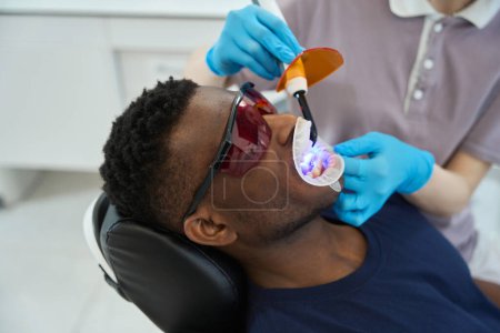 Téléchargez les photos : Jeune femme médecin dans des gants de protection debout près de l'homme et faire le nettoyage par ultrasons des dents dans la dentisterie - en image libre de droit