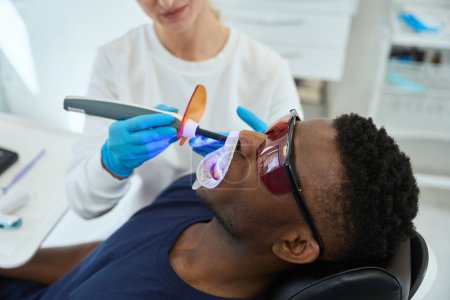 Téléchargez les photos : Infirmière dans des gants de protection debout près du patient et faisant le nettoyage ultrasonique des dents dans le bureau de dentisterie - en image libre de droit