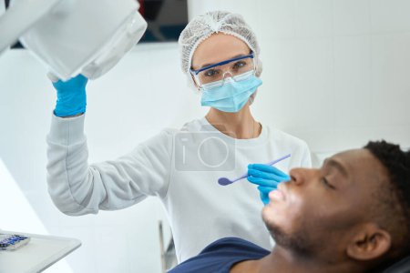 Téléchargez les photos : Femme en masque protecteur et gants debout près de l'homme et tenant miroir dentaire à l'hôpital - en image libre de droit