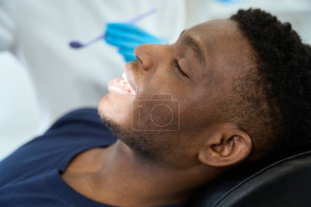 Téléchargez les photos : Jeune homme assis sur une chaise, femme médecin dans des gants de protection tenant miroir dentaire dans la clinique - en image libre de droit