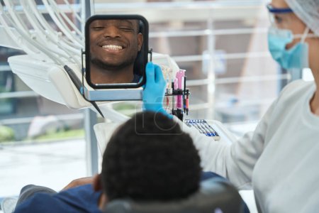 Photo for Dentist in uniform and mask and protective hat standing near man, holding mirror and talk with patient - Royalty Free Image