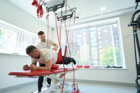 Foto de Paciente realizando tablón de antebrazo suspendido en gimnasio supervisado por fisioterapeuta calificado - Imagen libre de derechos