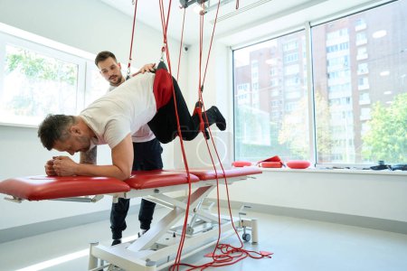 Photo for Adult patient doing suspended prone hip flexion under guidance of qualified kinesiotherapist - Royalty Free Image