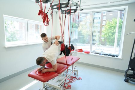 Foto de Varón haciendo flexión de cadera propensa suspendida con ayuda de entrenador de fitness profesional - Imagen libre de derechos