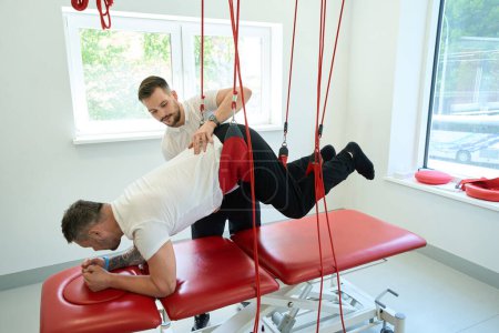 Foto de Fisioterapeuta enfocado que ayuda al paciente a realizar flexión de cadera propensa durante el entrenamiento de suspensión - Imagen libre de derechos