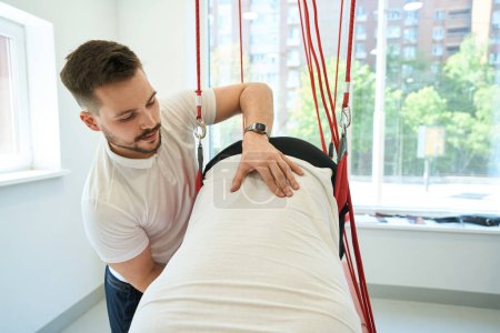 Photo for Kinesiotherapist assisting patient in performing exercise with suspended straps in rehabilitation center - Royalty Free Image