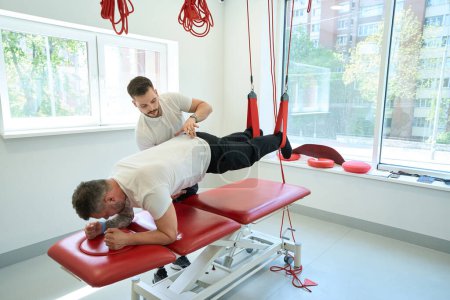 Téléchargez les photos : Physiatre concentré aidant le patient à faire un exercice de pontage sur un entraîneur de suspension - en image libre de droit