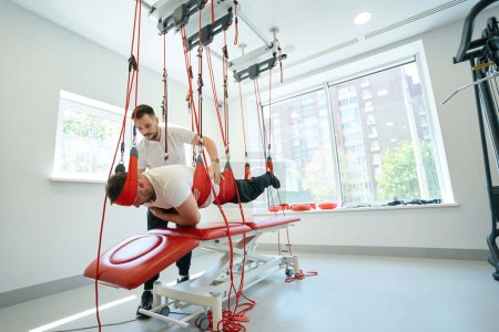 Téléchargez les photos : Exercice du client en position couchée sur un système d'entraînement basé sur la suspension assisté par un physiatre - en image libre de droit