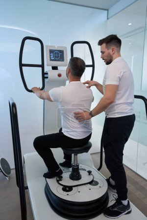 Téléchargez les photos : Homme faisant de l'exercice en position assise sur une plateforme motorisée multi-axes avec l'aide d'un physiothérapeute - en image libre de droit