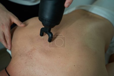Téléchargez les photos : Physiothérapeute qualifié desserrant les muscles paraspinaux serrés du patient avec un masseur percussif portatif - en image libre de droit