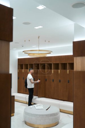Photo for Side view of man with backpack over his shoulder opening locker door in changing room - Royalty Free Image