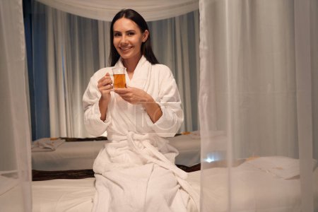 Foto de Alegre cliente de spa en albornoz sentado en el sofá de masaje sosteniendo la taza de bebida en sus manos - Imagen libre de derechos