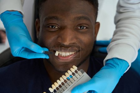 Foto de Enfermera en guantes protectores elige el color de los dientes de esmalte hombre antes de blanquear en la clínica - Imagen libre de derechos
