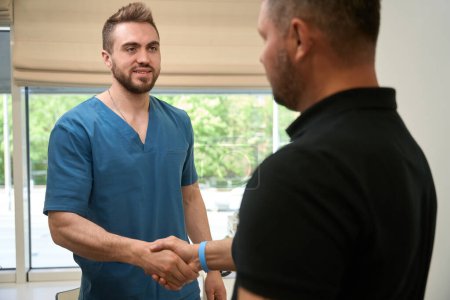 Téléchargez les photos : Souriant jeune médecin caucasien en bleus gommages serrant la main de l'homme adulte avant consultation - en image libre de droit