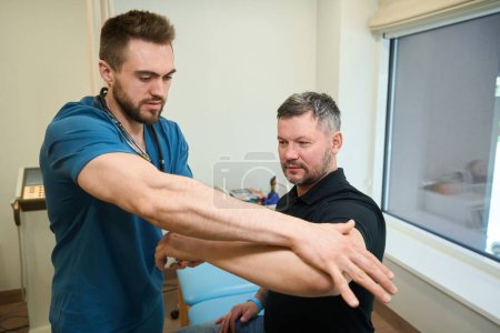 Photo for Adult man seated on examination couch while physiotherapist applying pressure to his forearm during neurological exam - Royalty Free Image