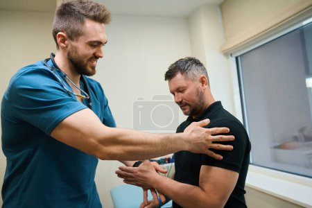Man sitting on examination couch while physical therapist assessing his arm muscle tone during physical exam