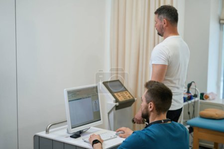 Téléchargez les photos : Médecin assis au bureau regardant l'écran de l'ordinateur pendant que le patient subit une analyse de la composition corporelle sur l'analyseur - en image libre de droit