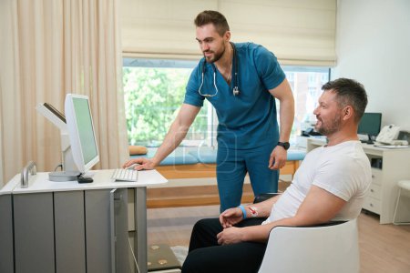 Foto de Paciente sonriente sentado en silla mientras el cardiólogo monitorea su rastreo de ECG en la pantalla de la computadora - Imagen libre de derechos