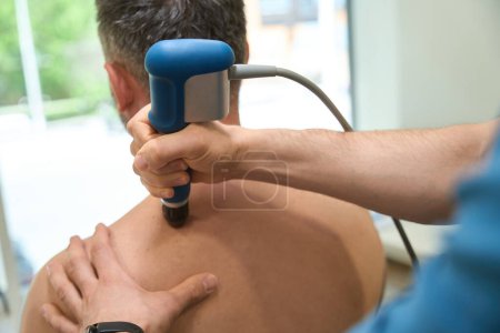 Foto de Foto recortada del fisioterapeuta que usa un dispositivo de terapia de ondas de choque extracorpóreas para aliviar el dolor de espalda superior en el hombre adulto - Imagen libre de derechos