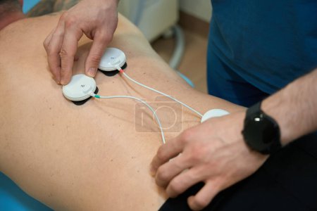 Foto de Foto recortada del médico colocando almohadillas de electrodo en la parte inferior de la espalda del cliente antes de la estimulación muscular eléctrica - Imagen libre de derechos