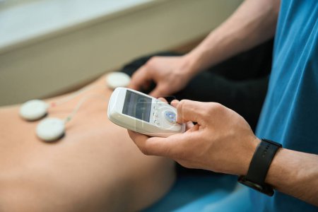 Photo for Cropped photo of physical therapist pressing button on electromyostimulation device while attaching electrode pads to client lower back - Royalty Free Image