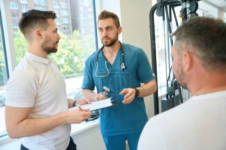 Foto de Neurólogo serio hablando con entrenador de rehabilitación frente al paciente en el gimnasio - Imagen libre de derechos