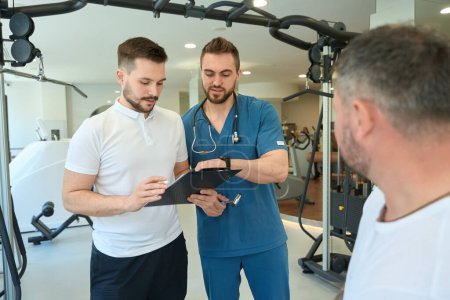 Téléchargez les photos : Physiatre ciblé montrant un plan de réadaptation physique à l'entraîneur de conditionnement physique en présence du client - en image libre de droit