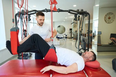 Téléchargez les photos : Homme adulte faisant flexion du genou couchée sur un entraîneur de suspension assisté par un physiothérapeute - en image libre de droit