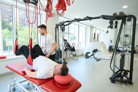 Photo for Adult person performing supine knee flexion exercise on suspension trainer assisted by fitness coach - Royalty Free Image