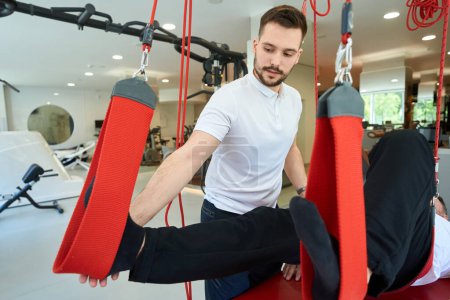 Téléchargez les photos : Physiatre expérimenté aidant le client à faire un exercice de flexion du genou couché sur un entraîneur de suspension - en image libre de droit