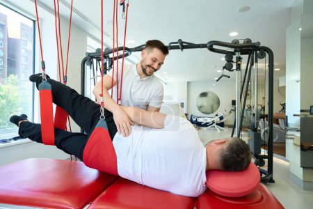 Photo for Client performing side-lying hip abduction on suspension trainer assisted by friendly gym instructor - Royalty Free Image