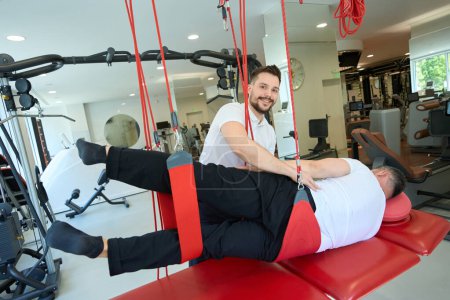 Photo for Cheerful physiatrist assisting patient in doing side-lying hip abduction on suspension trainer - Royalty Free Image
