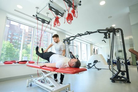 Photo for Male doing supine bridging with rotation on suspension trainer with help of fitness coach - Royalty Free Image
