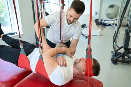 Photo for Rehabilitation center patient performing activation of trunk muscles on suspension trainer with help of kinesiologist - Royalty Free Image