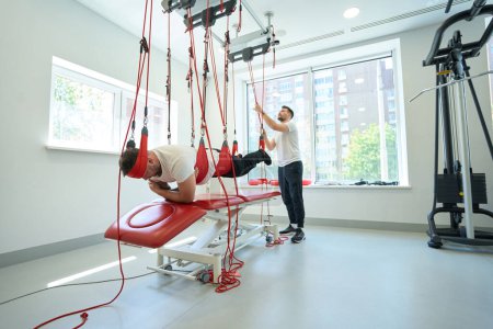 Foto de Paciente adulto que realiza activación muscular del tronco en sistema de entrenamiento basado en suspensión asistido por fisioterapeuta - Imagen libre de derechos