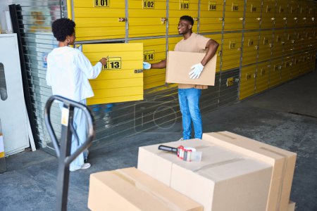 Foto de Hombre afroamericano cargando una caja de cartón en una unidad de almacenamiento, junto a una mujer gerente con una tableta - Imagen libre de derechos