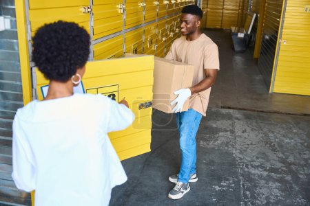 Photo for Young man loads a cardboard box into a storage unit, next to a woman manager with a tablet - Royalty Free Image