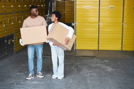 Téléchargez les photos : Jeune couple afro-américain en vêtements décontractés debout dans un entrepôt avec des boîtes en carton dans leurs mains - en image libre de droit