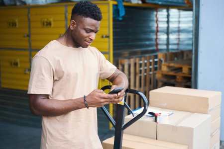 Photo for African American man stands with phone, leaning against cargo cart, on cart there are cardboard boxes and a tape dispenser - Royalty Free Image