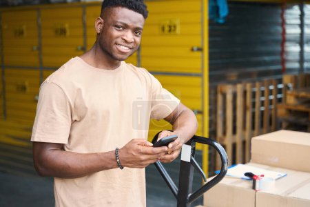 Photo for African American guy stands with phone, leaning against cargo cart, on the cart are cardboard boxes and a tape dispenser - Royalty Free Image