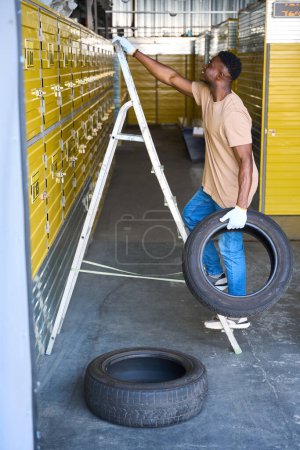 Téléchargez les photos : Un type grimpe un escabeau avec un pneu de voiture à la main, des conteneurs dans un entrepôt jaune - en image libre de droit