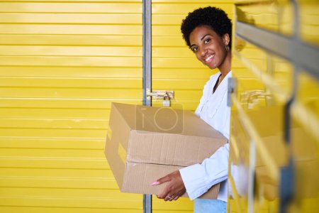 Téléchargez les photos : Jeune femme souriante debout dans un entrepôt avec boîte en carton, conteneurs dans un entrepôt jaune - en image libre de droit