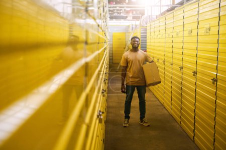 Téléchargez les photos : Afro-Américain en jeans se tient avec une boîte en carton près des conteneurs pour stocker des choses, les cellules sont numérotées - en image libre de droit