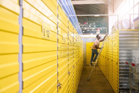 Foto de Un tipo en jeans se para en una escalera cerca de contenedores para almacenar cosas, las celdas están numeradas - Imagen libre de derechos