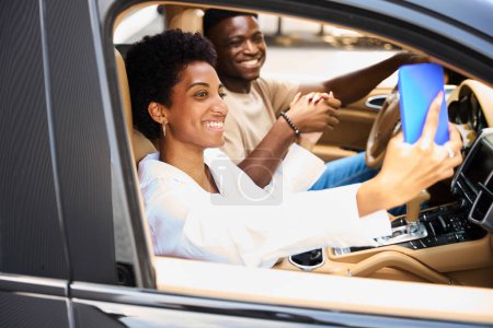 Téléchargez les photos : Joyeux couple afro-américain est assis dans une voiture confortable, la femme prend un selfie - en image libre de droit