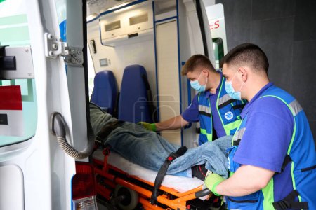 Foto de Hombres con máscaras protectoras descargan a un paciente de una ambulancia, el paciente se fija en una camilla especial - Imagen libre de derechos